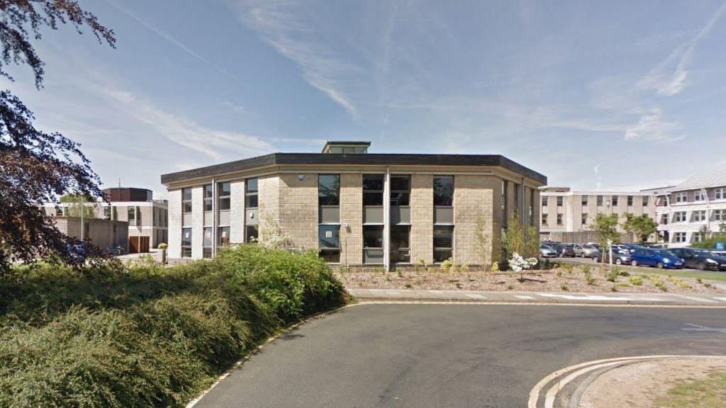 A google maps image of an octagonal brick building on a university campus with a road leading up to it and a carpark between other campus buildings