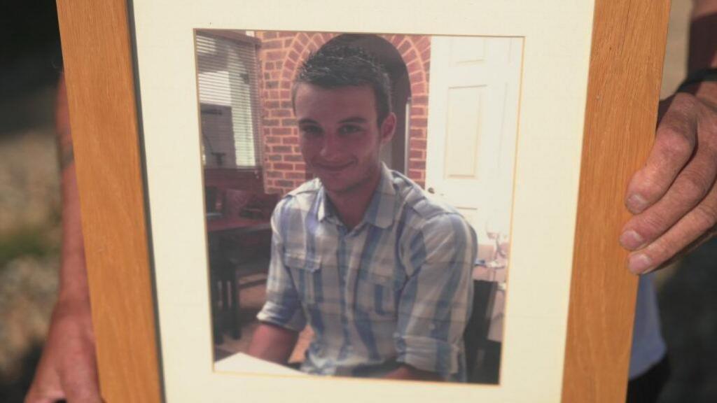 A framed picture of Daniele Polito, who is wearing a blue checked shirt with the sleeves rolled up. He is sat smiling at a table. A pair of hands can be seen either side of the wooden picture frame, holding it up
