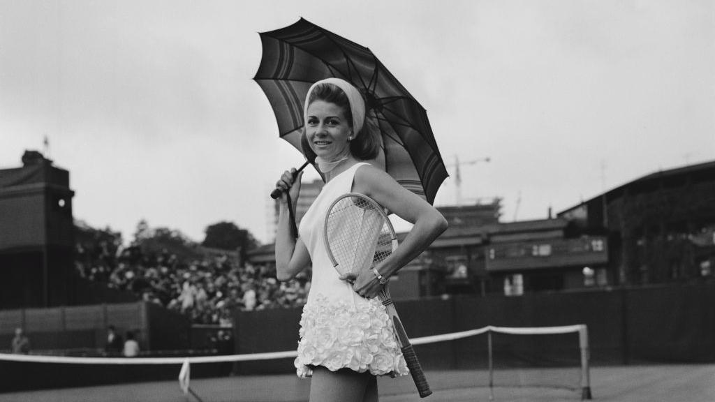 Pericoli is wearing a white dress frilled with lace roses on the skirt, holding a racquet in one hand and a parasol in the other.