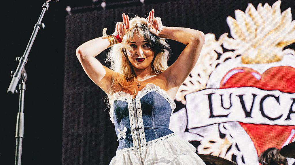 Luvcat performs onstage during Tramlines at Hillsborough Park. She puts her fingers on her head like cat ears.