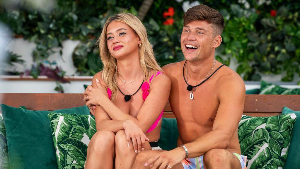 Former US Love Island contestants Kaylor Martin and Aaron Evans are sat beside each other on a sofa, which has green cushions. The pair are dressed in swimming costumes.