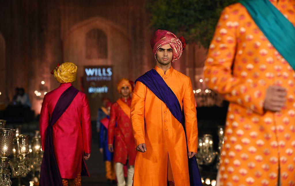 Models wear clothes designed by Rohit Bal during the final of Indian Fashion Week in 2015. A man in the centre of the image is wearing an orange sherwani, a blue sash, and red turban.