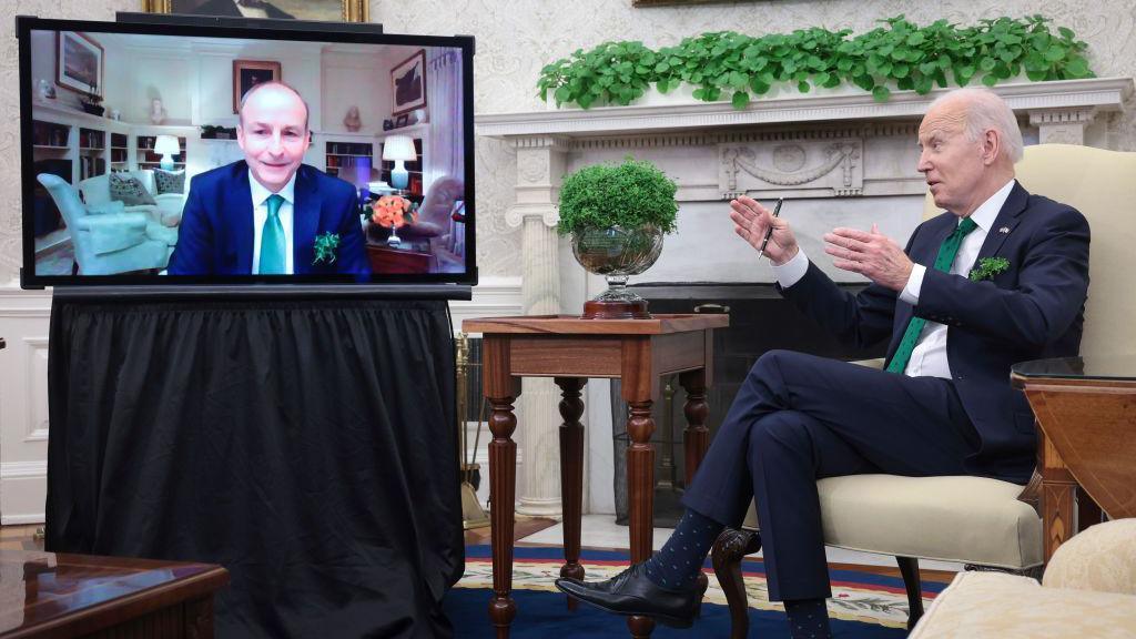 Micheál Martin pictured on a TV screen in the White House during a video call with former US President Joe Biden in March 2022.  Both men are wearing navy suits, white shirts and green ties.  A bowl of shamrock is on a table in front of Biden. 