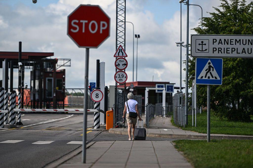 A woman pulling a small suitcase approaches a road crossing. There is a barrier, to her left and gates and fences in front of her. There is a red stop sign in the foreground.