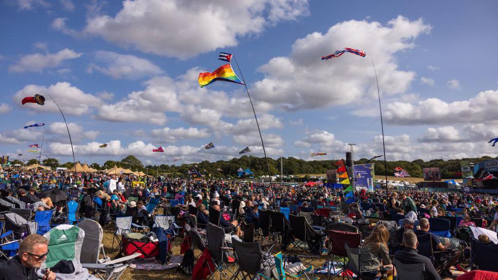 A wide view of a festival during the day, with people seated in camping chairs and colourful flags flying overhead.