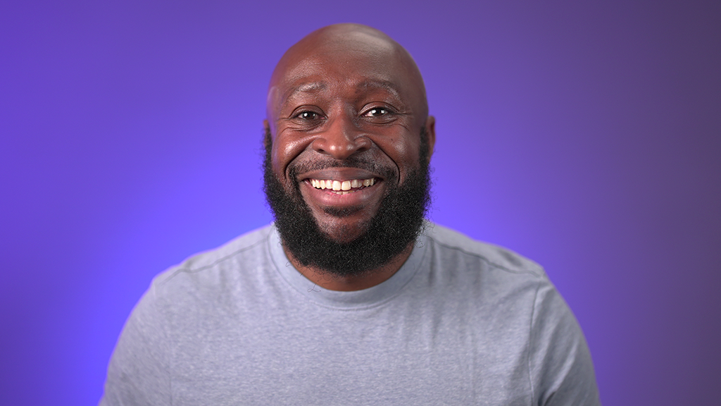 Riley wears a grey shirt and smiles at the camera in front of a blue background.
