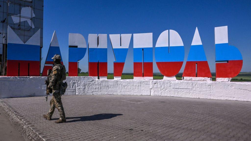 A uniformed Russian soldier walks in front of a sign that spells "Mariupol" in Cyrillic, with the letters painted the colours of the Russian flag.