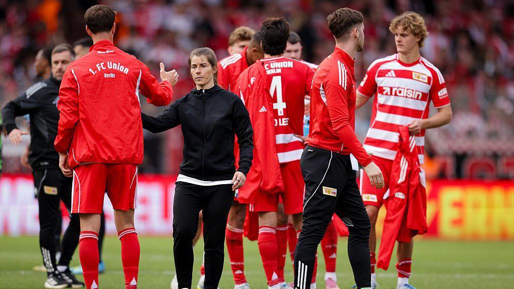 Marie-Louise Eta consoles her players as they lose her first game in charge of Union Berlin. She is wearing a black tracksuit. The players are wearing red and white hooped shirts with red shorts and socks.