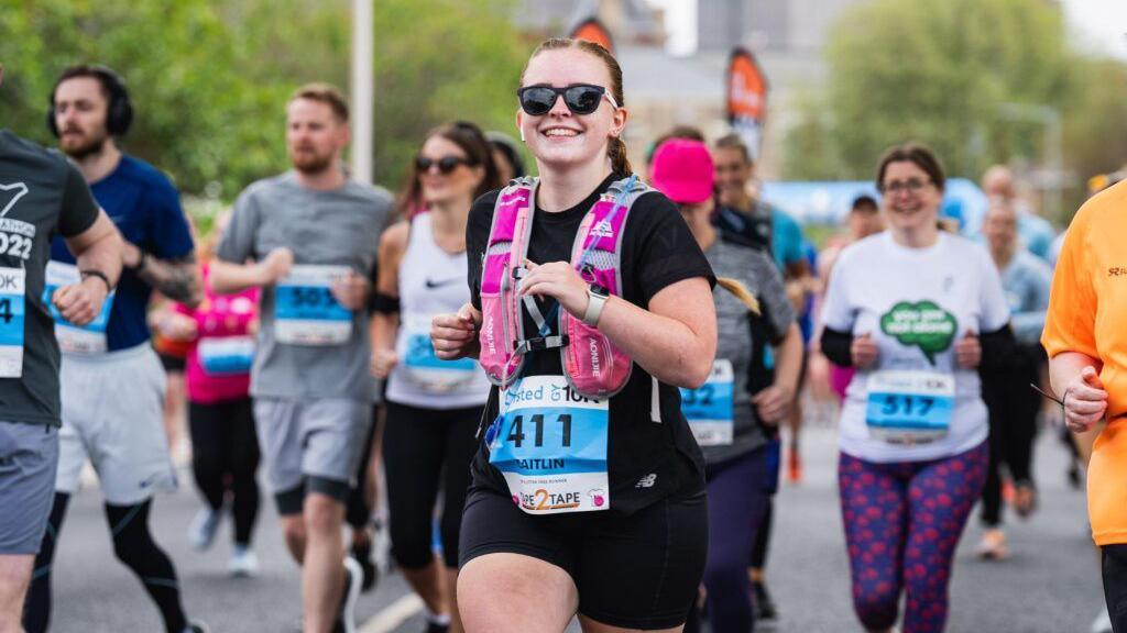 A runner wearing sunglasses, black t-shirt, black shorts and a pink hydration vest smiles into the camera as she takes part in the Grimsby 10K. A number of other runners are visible in the background.