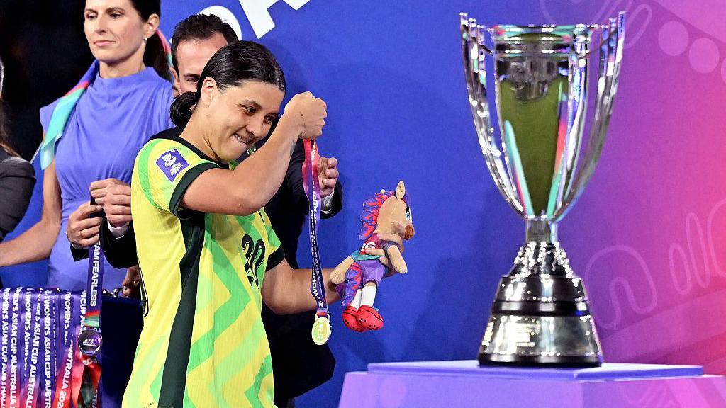 Sam Kerr takes off her medal as she passes the Asian Cup trophy