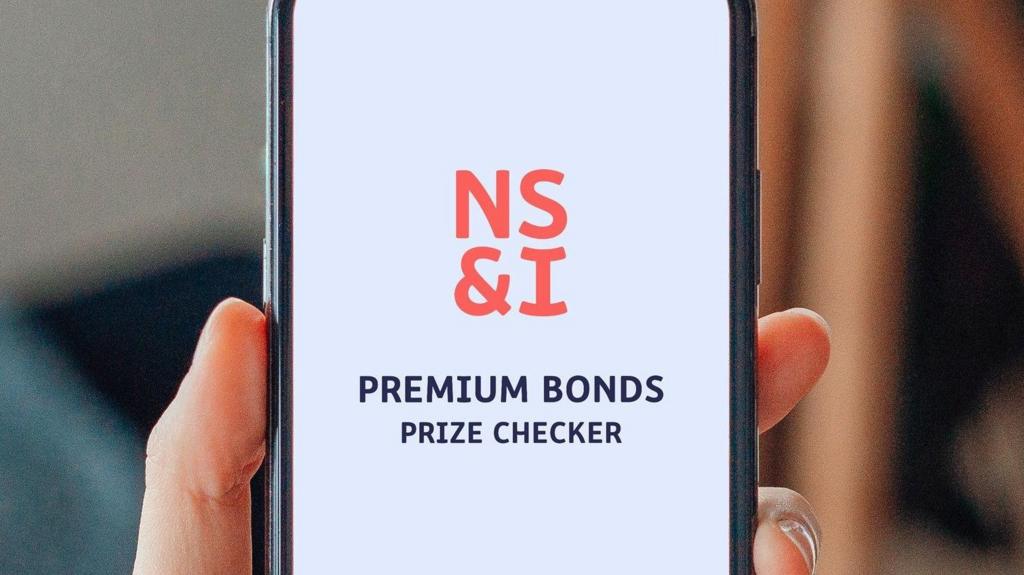 A hand holding up a phone. The screen shows the opening page of the NS&I app Premium Bonds prize checker app