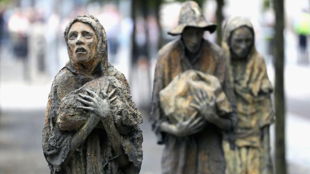 Bronze sculptures, three emaciated humans, gripping bags. 