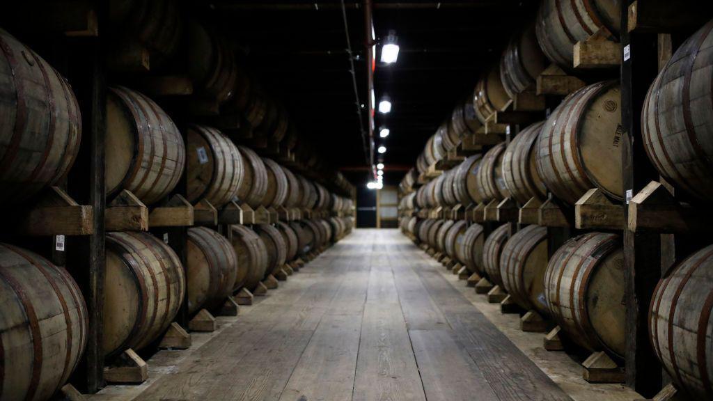 A long wooden hall is flanked by stacks of wooden bourbon barrels on either side, with industrial lights illuminating the way