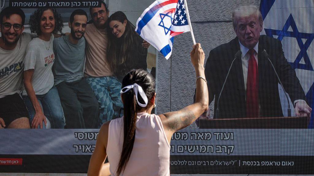 A woman waves Israeli and US flags while watching a screen that shows side-by-side footage of released Israeli hostages and US President Donald Trump