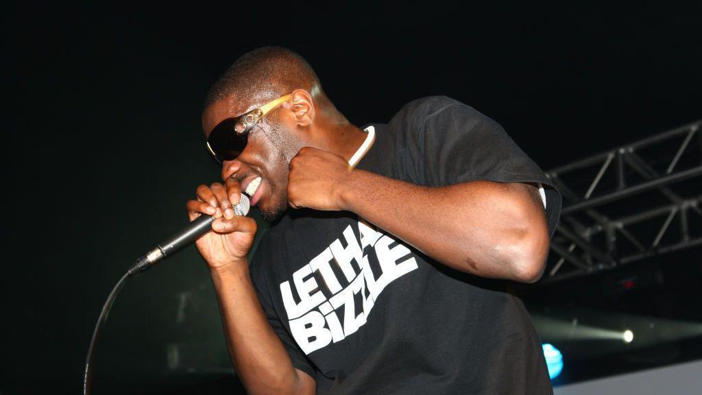 Lethal Bizzle, facing towards the left of the camera, holds a microphone to his mouth as he performs on stage while wearing a black t-shirt with his name written on it in white lettering