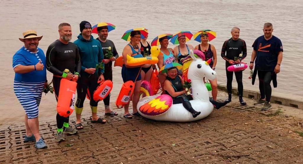 Twelve men and women stood in front of the Humber on the pavement wearing swimming uniform. Some of the swimmers have multi-coloured umbrella hats on their head. One swimmer is sat in a unicorn inflatable. 