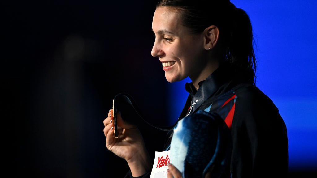 A young woman with dark hair tied back in a hair band. She wears a dark tracksuit and smiles at the cameras while holding her gold medal.