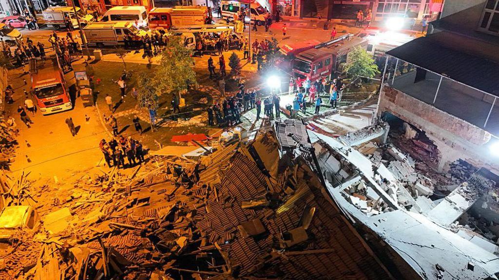 An aerial view of a collapsed building with emergency workers all around it. The picture is taken at night time and the debris is lit by strong spot lights.