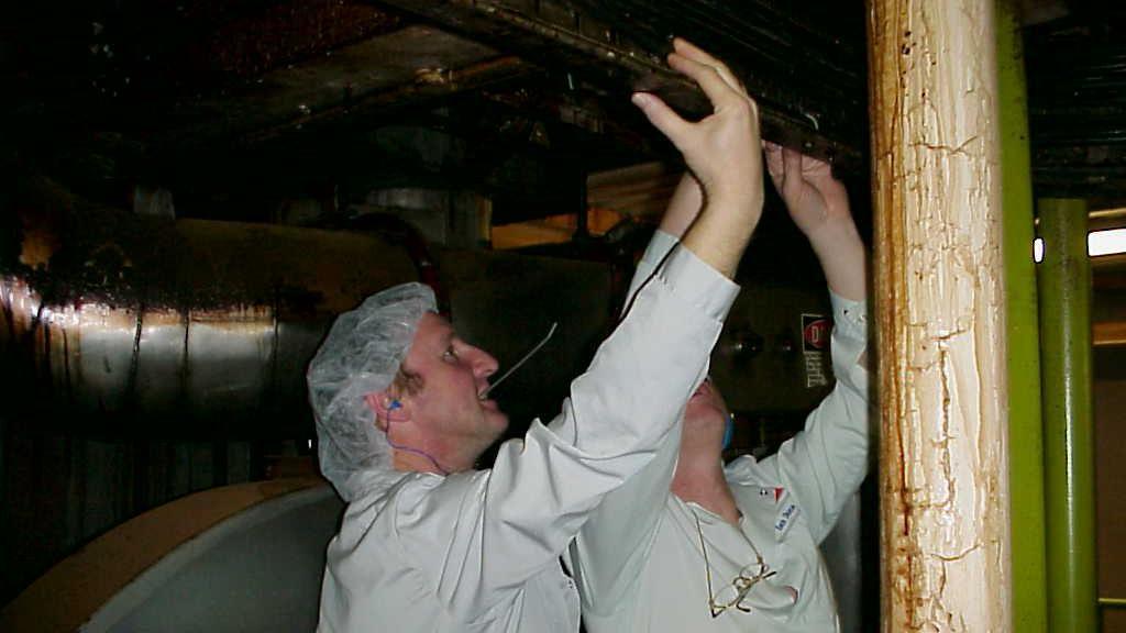 Two people dressed in white overalls and hair nets look at a pipe.