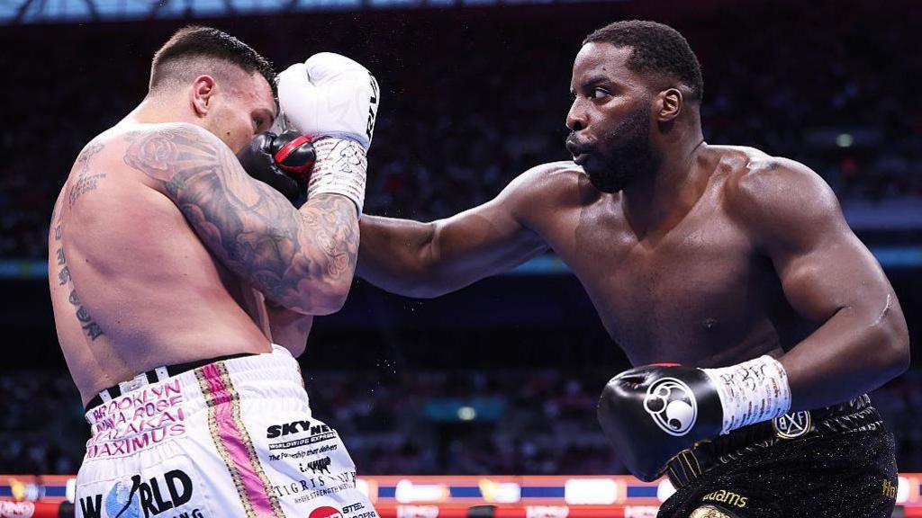 Two male boxers are fighting in a brightly lit stadium ring in front of a large crowd. The boxer on the right, Lawrence Okolie, is a tall, muscular black man wearing black and gold trunks and black gloves. He is throwing a powerful right-hand punch toward his opponent’s head. The boxer on the left, Krzysztof Glowacki, is a heavily tattooed, white man with close‑cropped hair, wearing white trunks with colourful writing and white gloves. He is hunched over, arms raised tightly in front of his face and body as he absorbs or blocks the punch. Their bodies are very close, with sweat visible, and the moment captured feels tense and explosive, as if in the middle of an intense exchange during a high‑level boxing match