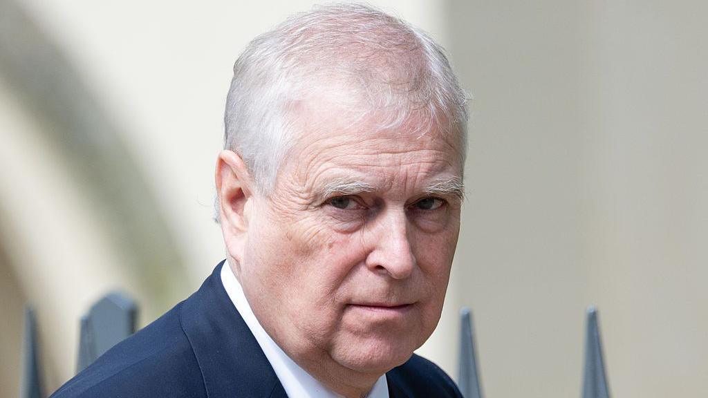 A close-up of Prince Andrew, who is outside in front of some railings - he is wearing a dark blue suit jacket and a white shirt which can just be seen and is looking at the camera with a neutral expression