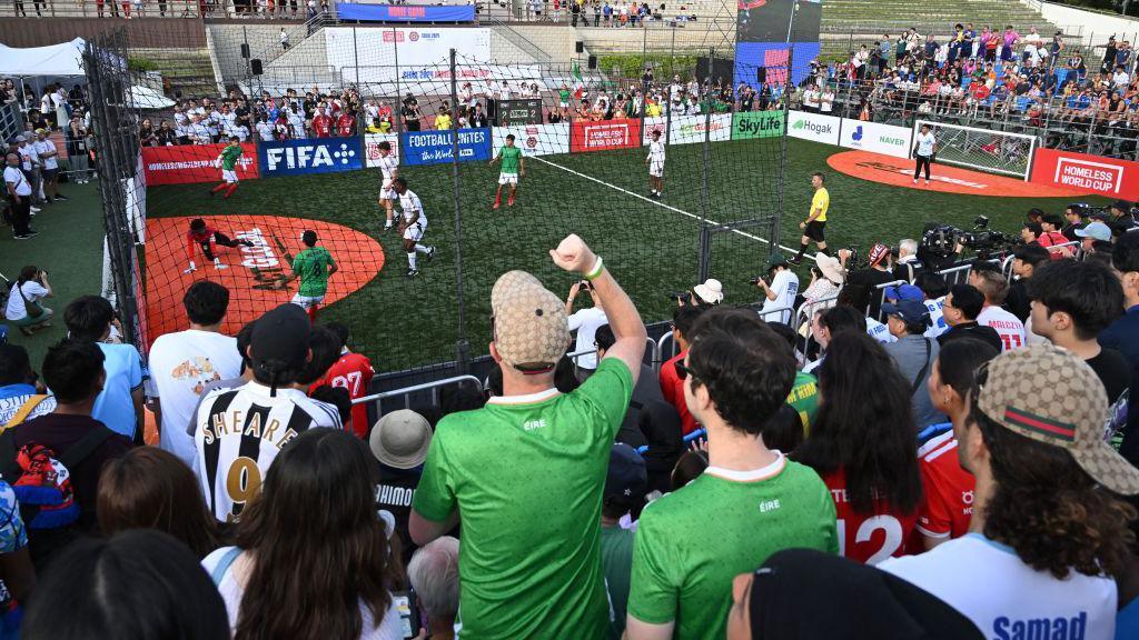 A crowd of people watching a football game on a small pitch. There are eight players on the pitch, some wearing green, and some wearing white.
