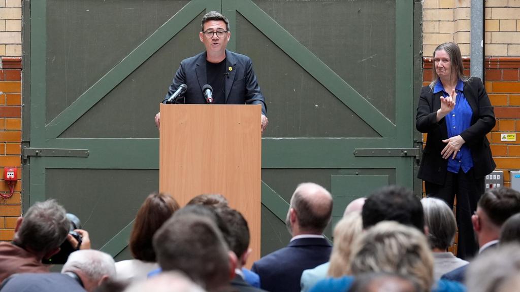 Burnham speaks at a podium with a sign language interpreter to his side and an audience in the foreground.