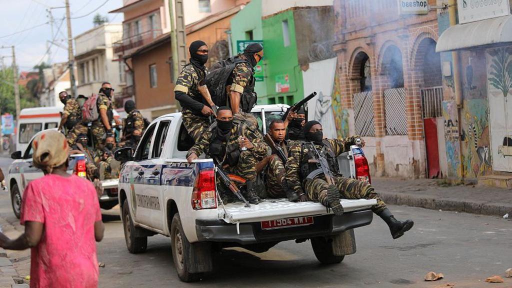 Armed members of the security forces in military fatigues can be seen in the back of an open van driving along a road. A second vehicle with security personnel can also be seen in front of them on the road.