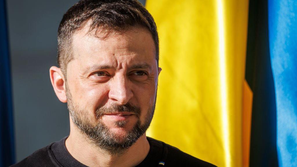 Volodomyr Zelensky, who has dark brown hair and facial hair, stands in front of a yellow and blue Ukraine flag.