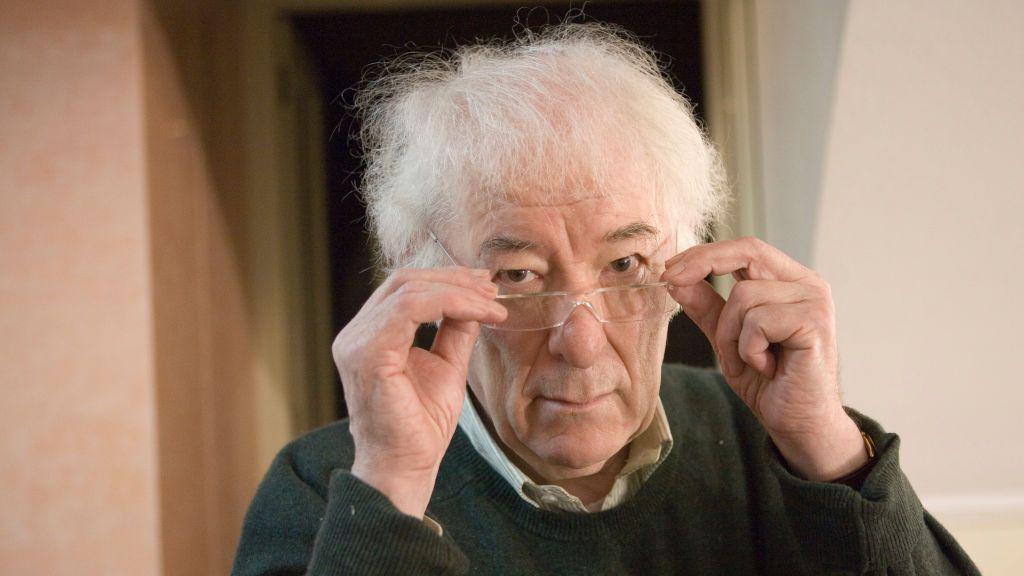 A man with white fluffy hair holds his glasses on the end of his nose. He looks at the camera with a neutral expression.