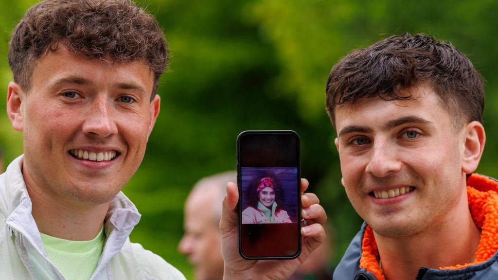 Two men are side by side. The man on the left is holding up a phone between the pair. On the mobile phone, is a picture of a young woman, in her 20s, with curly dark hair and a red hairband. The man on the left has short curly hair and is wearing a green t-shirt and light-coloured waterproof running jacket. The man on the right has short dark hair, and is wearing a dark rain jacket with orange padding. They are both smiling for the camera