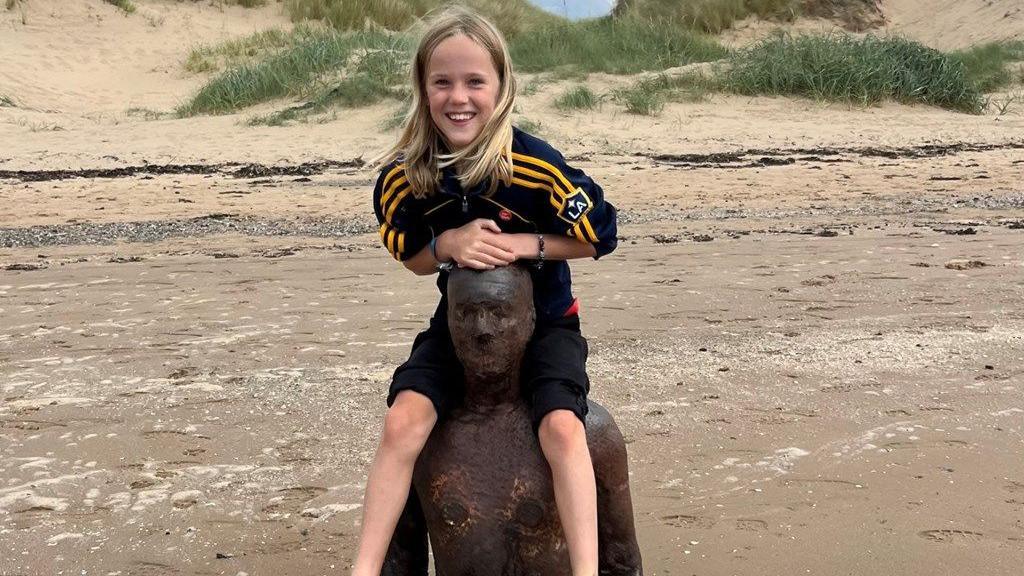 Evan Rothwell has long blonde hair and smiles at the camera as he sits on the shoulders of an Iron Man statue on Crosby beach.