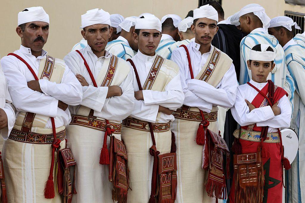 Men and boys dressed in white and red Amazigh clothing stand in a row.
