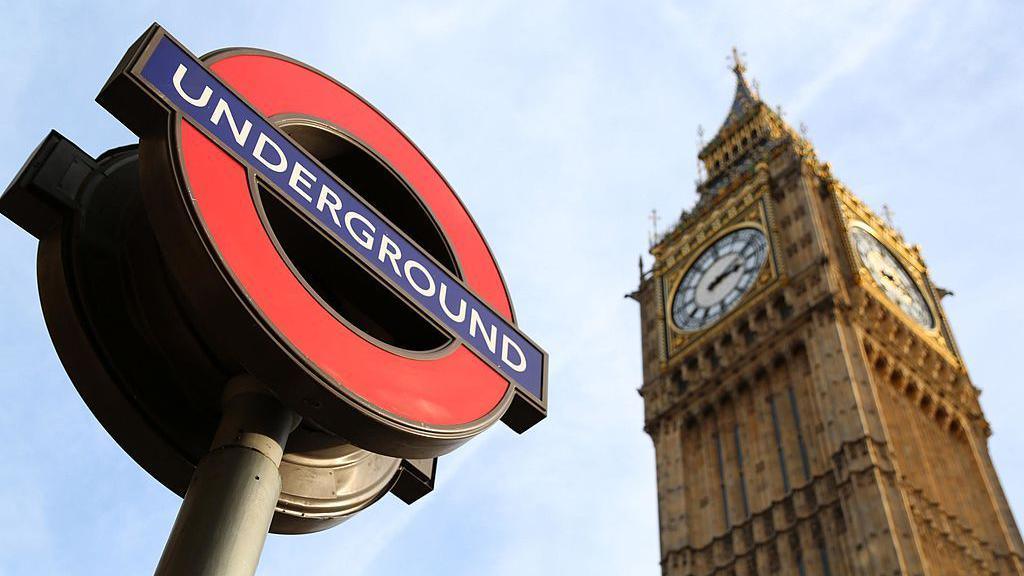 A signpost displaying the London Underground roundel logo, with the Big Ben clock tower on the Houses of Parliament in the background.