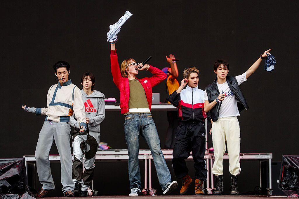 Five boyband members wearing jackets and jeans performing on a stage
