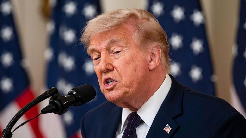 Donald Trump mid-speech speaking into two thin black mics, several US flags on poles behind him in the blurred background