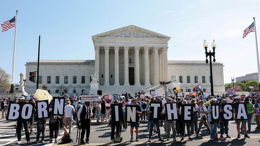 A crowd of people holding letters that spell out 'born in the USA' stand in front of a large white pillared building