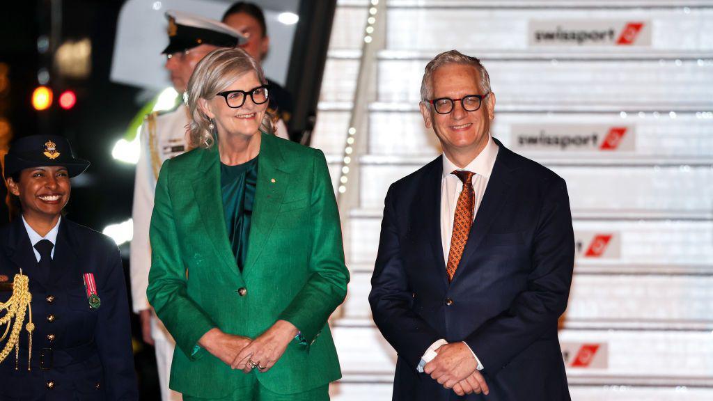 Sam Mostyn wears a green blazer and stands next to her husband Simeon Beckett. The pair stand in front of the airplane smiling.