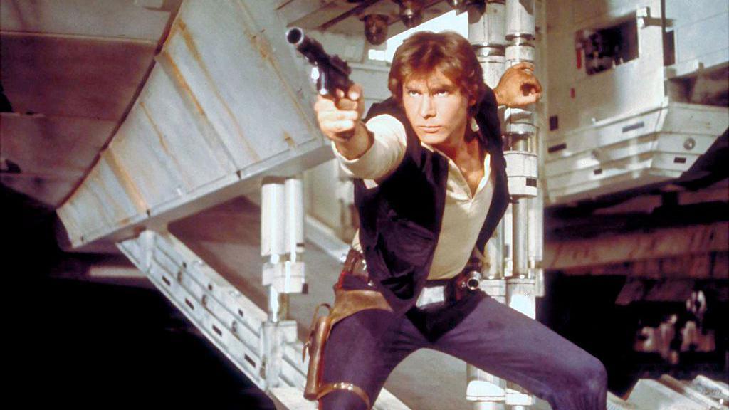 Actor Harrison Ford, as Hans Solo, on the set of Star Wars: Episode IV - A New Hope. He is in a fighting stance and holds up a futuristic gun.