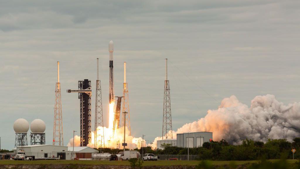 A slender rocket launches into space with a large plume of smoke. In front of the rocket is a large warehouse. 