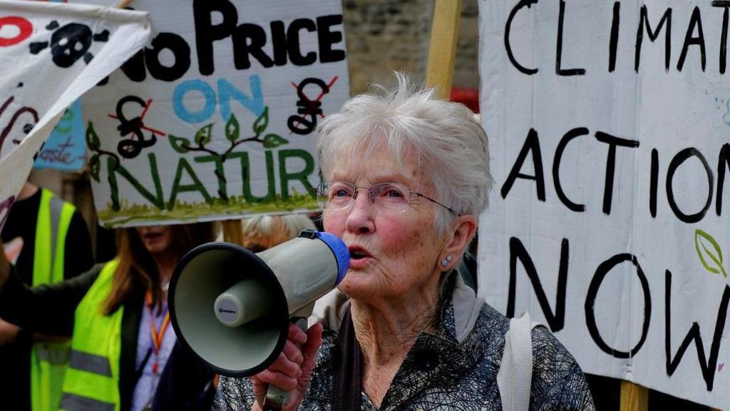 Peggy Seeger campaigning near the Carfax Tower in Oxford with a megaphone. Other people can be seen behind her holding placards with environmental slogans.