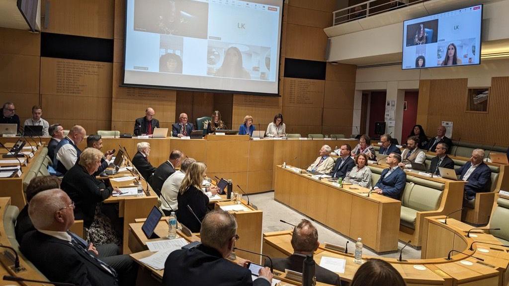 The inside of a packed council meeting shows councillors sat at desks in a hall while other attend via large screens hanging overhead.