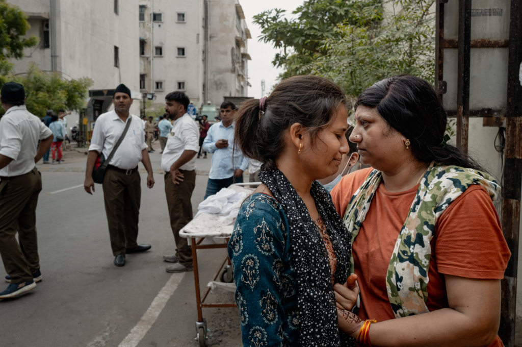 Two women, one wearing blue and one wearing orange, hold each other in the street. Men stand in the background, a trolley sits just behind them.