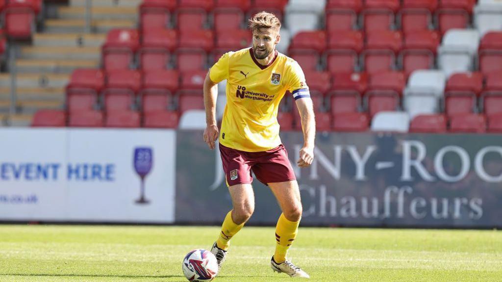 Captain Jon Gutherie in a yellow football top and burgundy shorts  looks to pass the football at a stadium. 