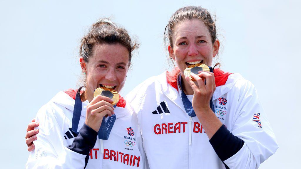 Two women in white sports clothes bite gold medals they have won in a rowing race. 