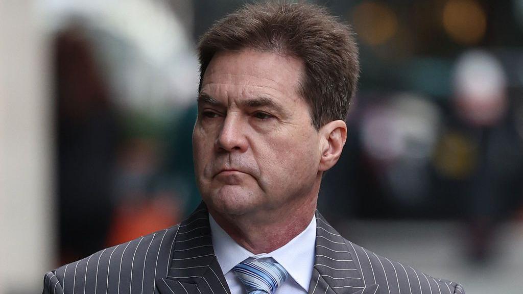 Craig Wright, a white man in his fifties with short brown hair. He is wearing a sharp pinstripe suit with a striped tie and white shirt.