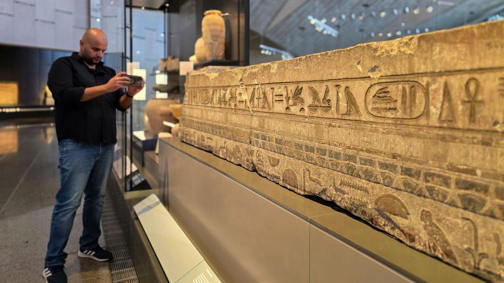 A visitor taking a photograph of an ancient stone wall covered in hieroglyphs