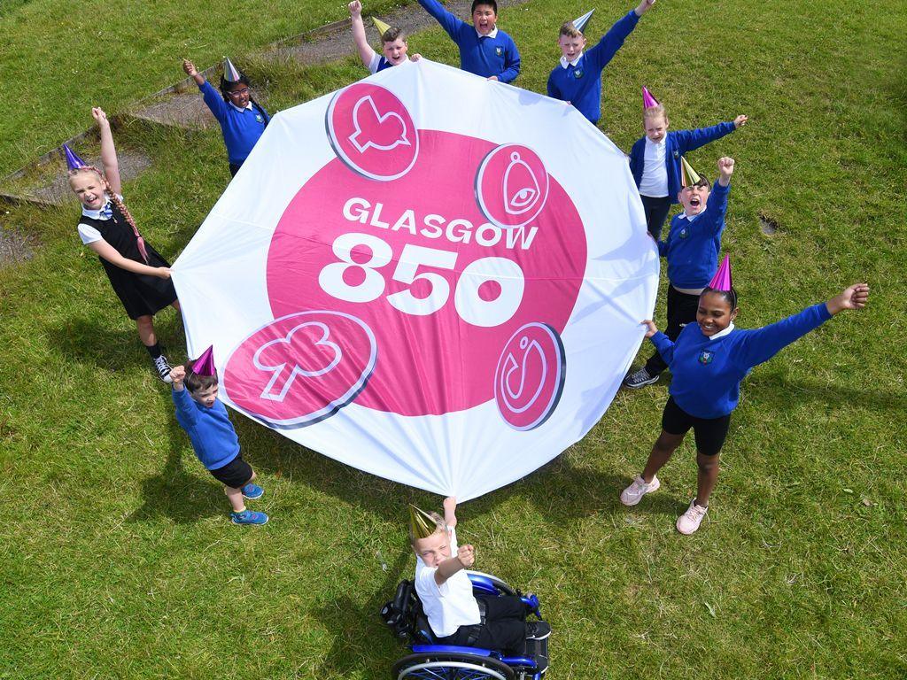 Children wearing uniforms from Saracen Primary School in Possilpark and party hats hold white and pink banner showing the Glasgow 850 branding badge images