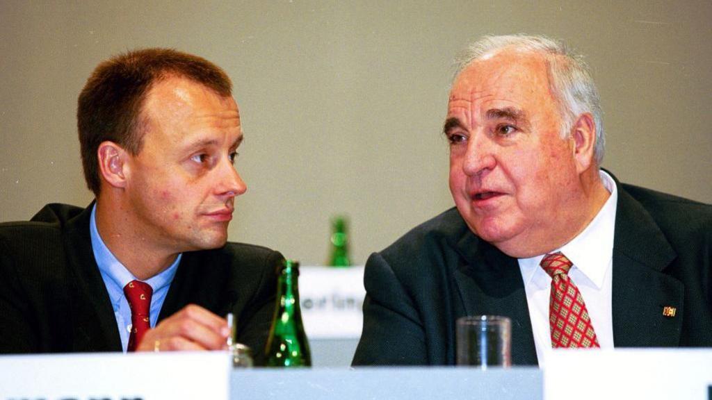 Friedrich Merz in a blue tie on the left listens to the older Helmut Kohl talking 