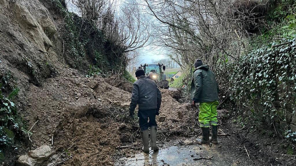 Two men and a tractor clearing soil and debris from a road after a landslip.